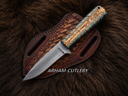 A fixed-blade knife with a silver steel blade and a carved wood handle wrapped in blue cord, resting diagonally on a brown leather sheath over a dark brown fur background. The text 'ARHAM CUTLERY' is overlaid at the bottom.