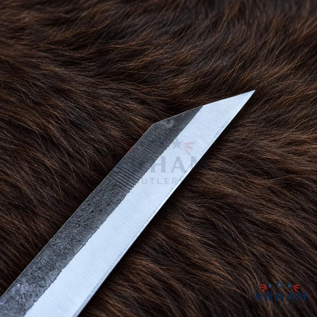 A close-up of a metal knife blade resting diagonally on a dark brown fur background. The blade has a brushed silver finish on one side and a textured dark gray finish on the other, with a faint watermark reading 'SHAN.