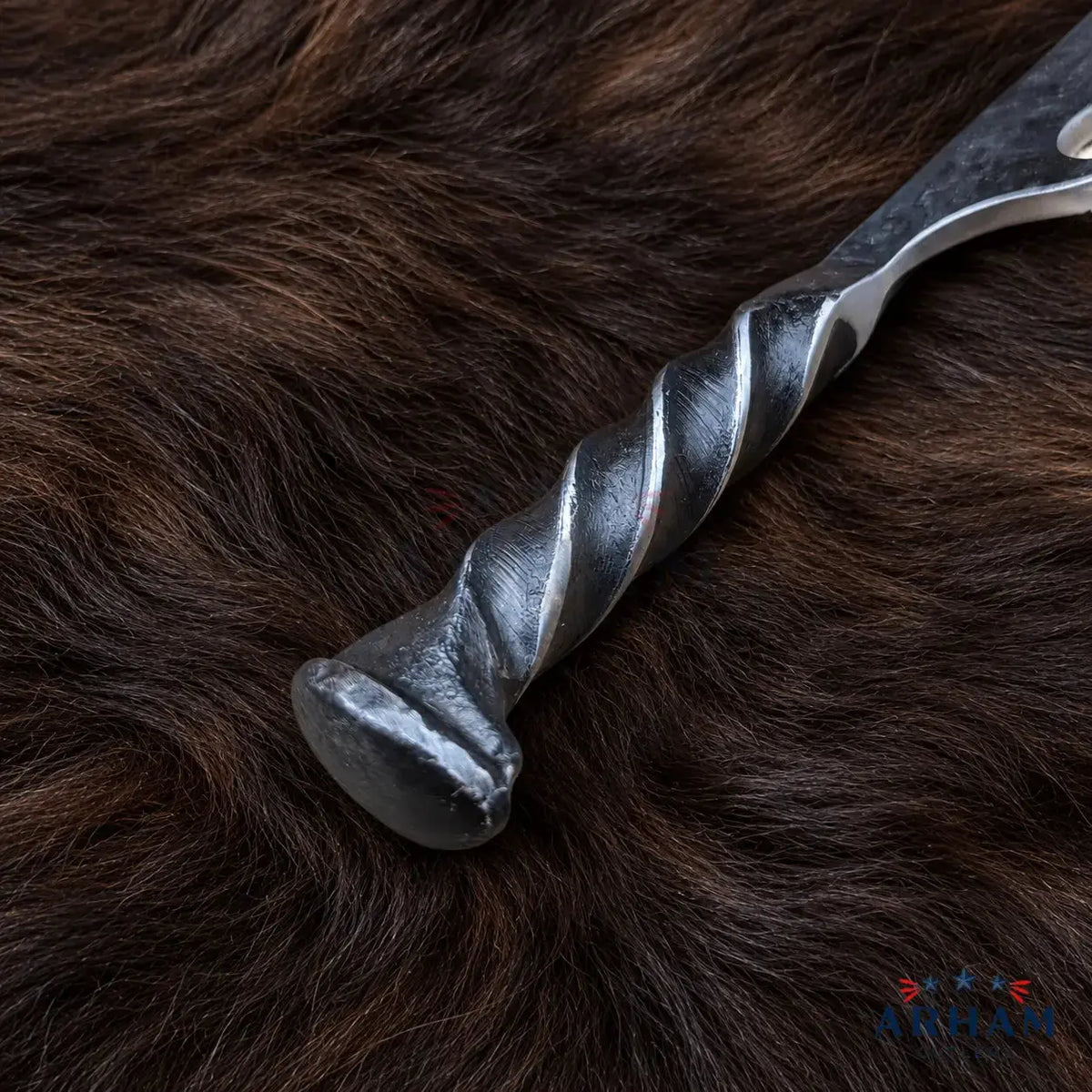 A close-up of a dark metal dagger or knife handle with a twisted spiral grip, resting on a brown fur background. The brand name 'Arkan' is visible as a watermark in the bottom right corner.