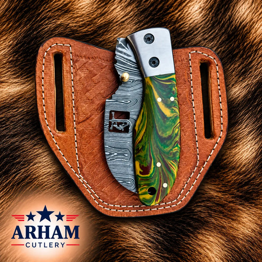 A folding pocket knife with a green and yellow marbled handle and a patterned steel blade, displayed in a brown leather sheath on a brown fur background. The ARHAM CUTLERY logo is visible in the bottom-left corner.