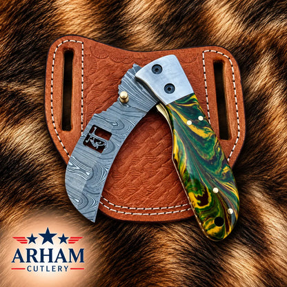 A folding knife with a silver Damascus steel blade and a green, yellow, and black marbled handle, displayed on a brown leather sheath over a brown fur background. The 'ARHAM CUTLERY' logo is visible in the bottom-left corner.