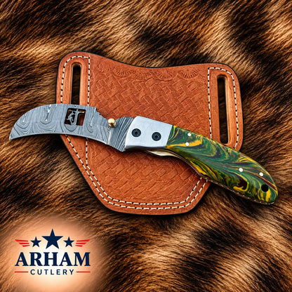 A folding pocket knife with a patterned steel blade and a green marbled handle, displayed on a brown leather sheath over a brown animal fur background. The ARHAM CUTLERY logo is visible in the bottom-left corner.