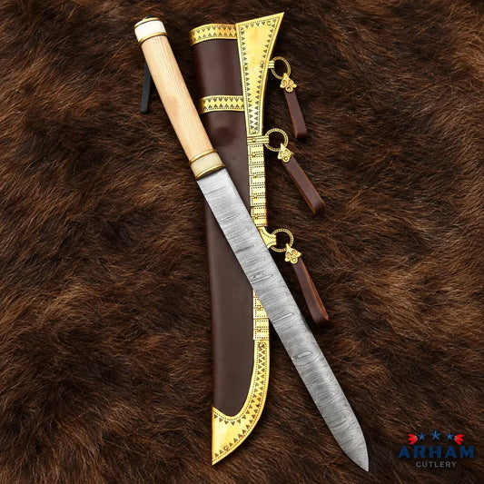 A Viking-style knife with a patterned steel blade, gold-toned metal fittings, and a light wood handle, displayed next to its brown leather sheath with gold trim, all laid on a brown fur background. The Arham Cutlery logo is visible in the bottom right corner.