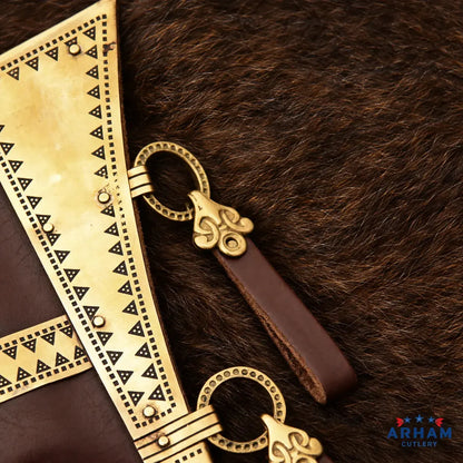 Close-up of a leather belt with an ornate brass-toned metal buckle and decorative triangular patterned plates, laid on dark brown fur. The ARHAM CUTLERY logo is visible in the bottom right corner.