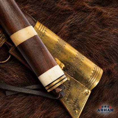A close-up of a decorative brass and wood object with carved geometric detailing, resting on dark brown fur. The item has a dark brown wooden handle with cream and black bands, and a polished brass body with engraved patterns.