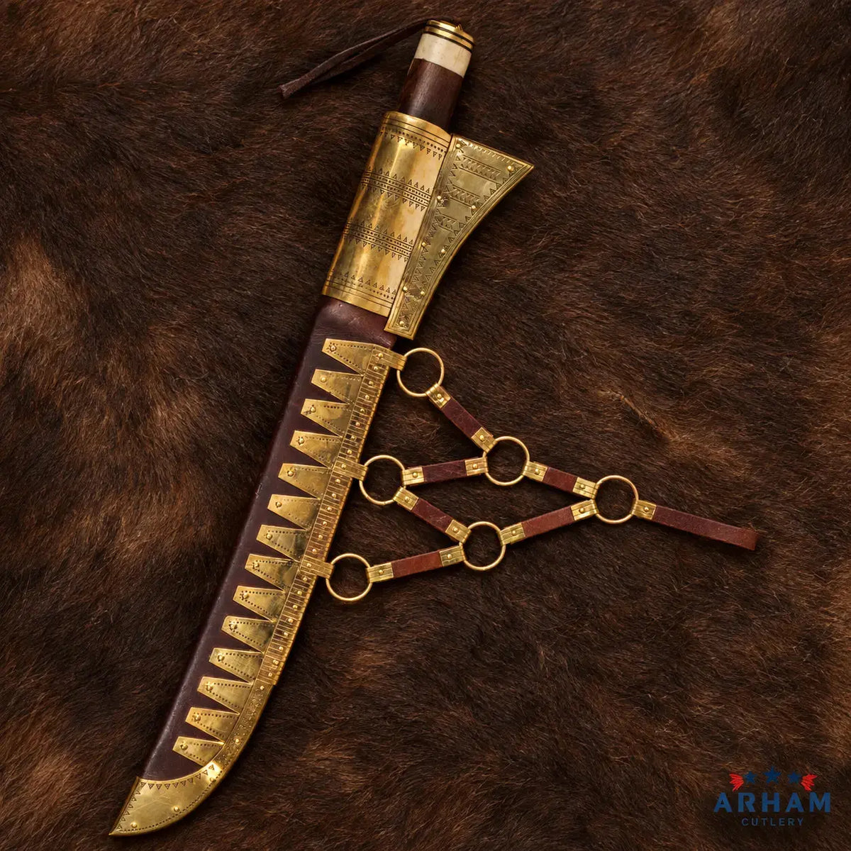 A decorative curved knife or dagger shown with its brown leather sheath on a brown fur background. The blade and sheath are adorned with ornate gold-toned metal plates, studs, and rings, while the handle is wrapped in dark brown and cream-colored material.