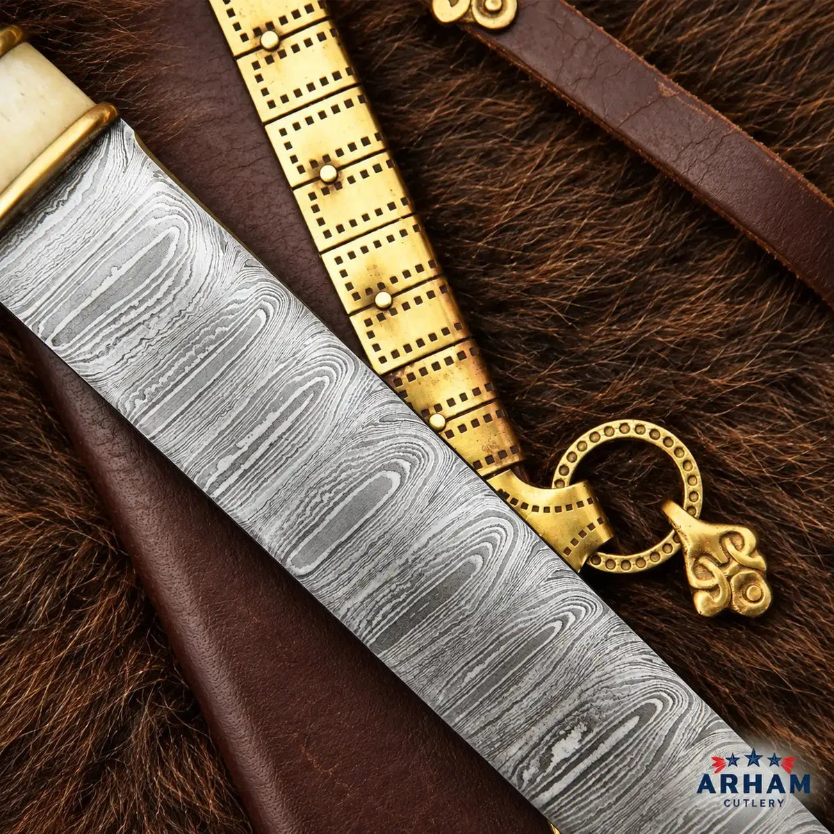 A close-up of a decorative dagger or sword with a patterned steel blade, shown resting on brown leather and dark fur alongside a gold-toned belt and a brass ring ornament. The ARHAM CUTLERY logo appears in the bottom right corner.