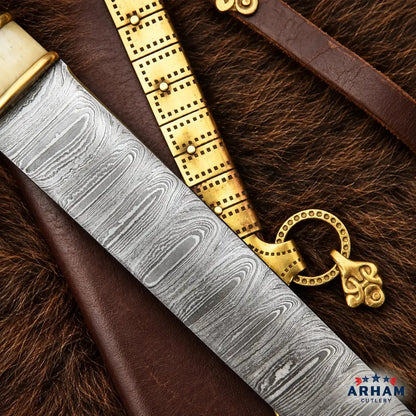 A close-up of a decorative dagger or sword with a patterned steel blade, shown resting on brown leather and dark fur alongside a gold-toned belt and a brass ring ornament. The ARHAM CUTLERY logo appears in the bottom right corner.
